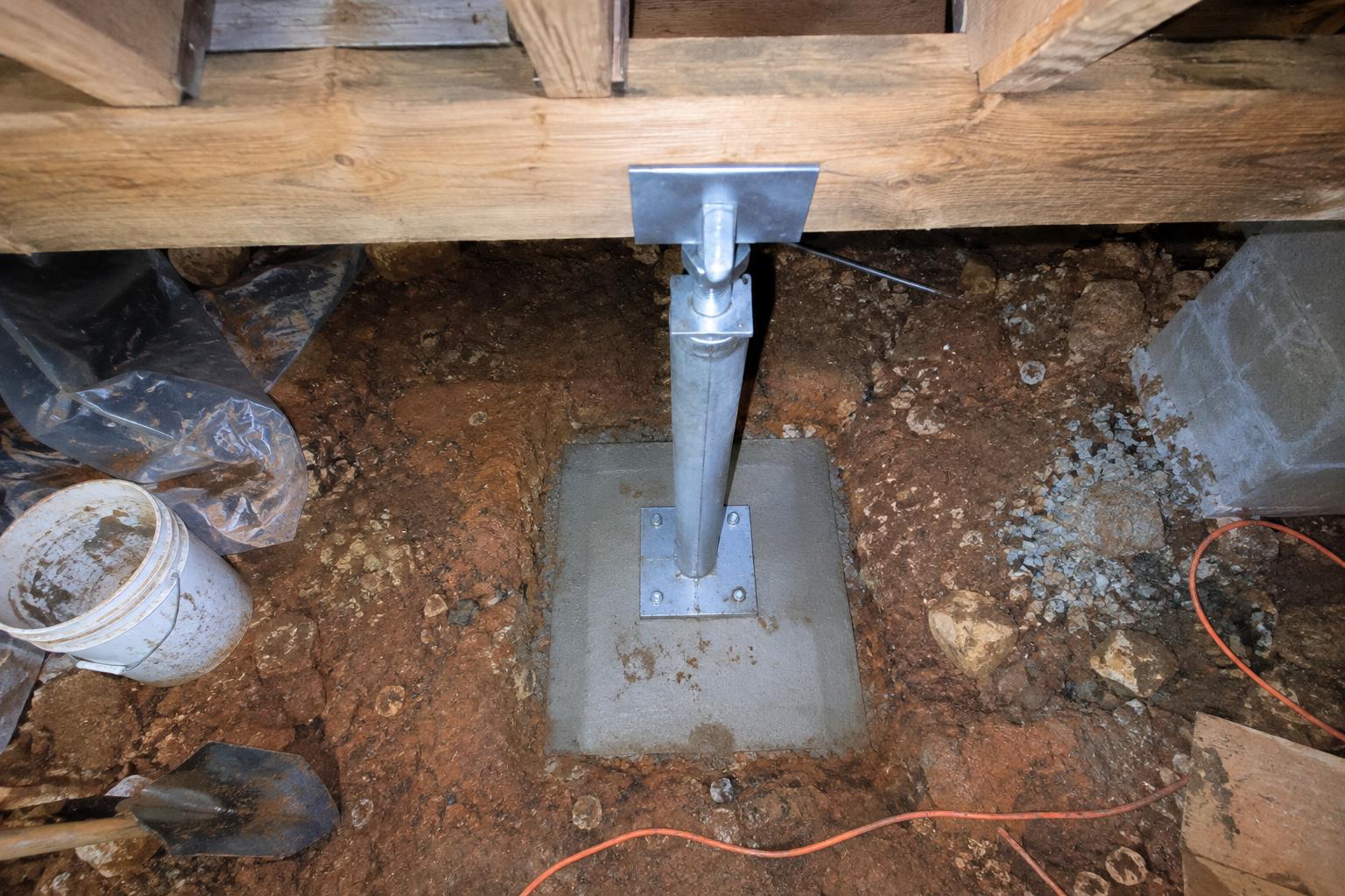 Adjustable steel support post set on a poured concrete pad under a wood main beam in a low crawl space, with floor joists above