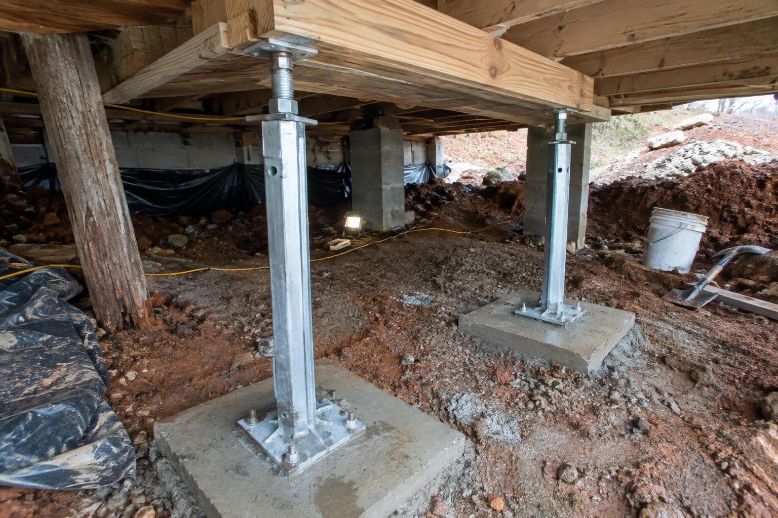 Two adjustable steel support jacks on poured concrete footing pads holding up a sistered wood floor joist in a low crawl space with damp dirt below.